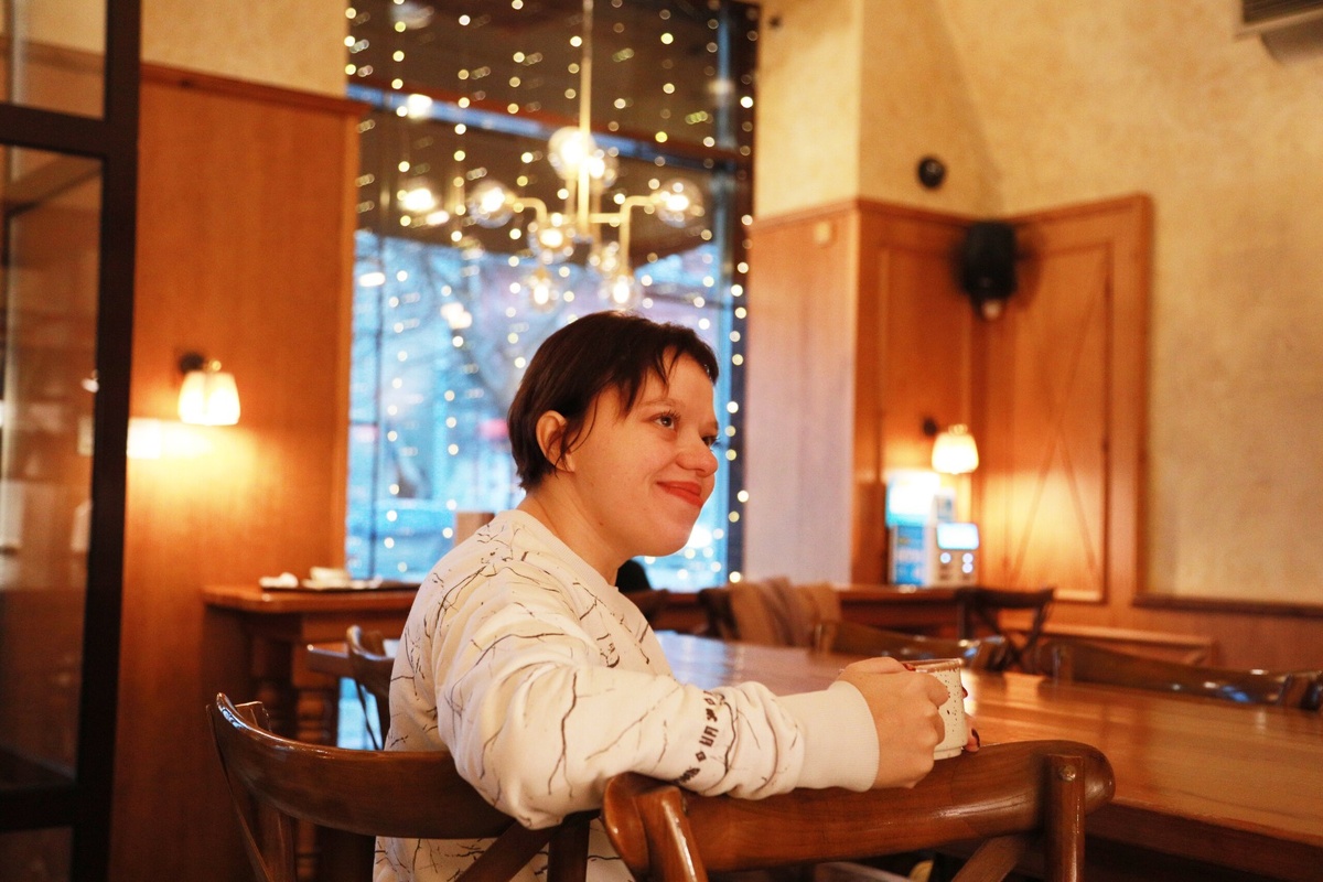 Тифлокомментарий. Ольга в кафе. Она сидит на высоком стуле за столом. На ней белый вязаный джемпер, в руках чашка кофе. На заднем плане окно с подсветкой.