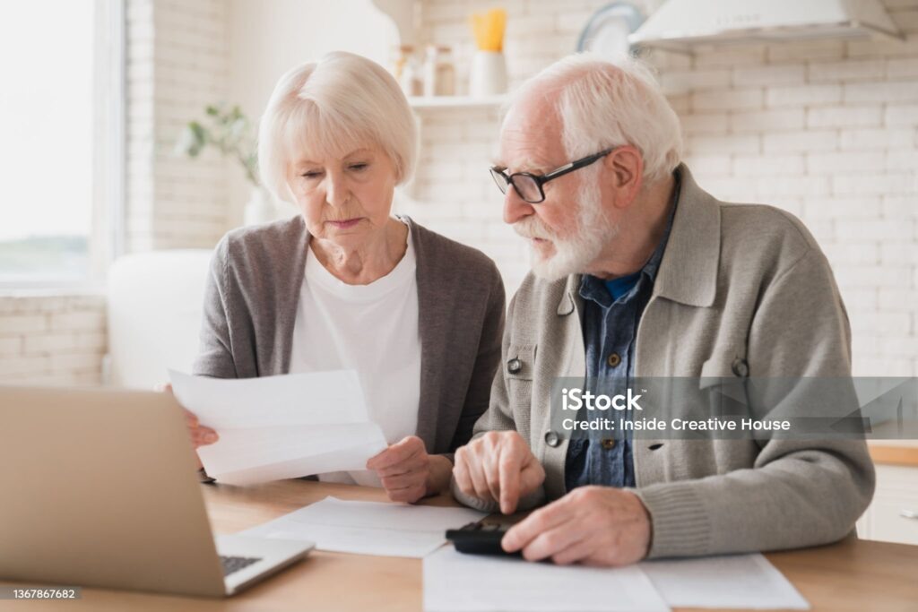    Serious caucasian old elderly senior couple grandparents family counting funds on calculator, doing paperwork, savings, paying domestic bills, mortgage loan, pension at home using laptop. Редакция газеты