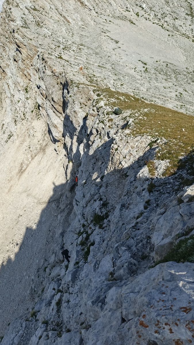 Перевал Дзоу в том месте, где лежит зелёный рюкзак. Прямо под ним стоит мой рюкзак – с ковриком в оранжевом чехле. К моему рюкзаку идёт мой напарник – по тропе, что под скалами. Фото моё. Август 2025.