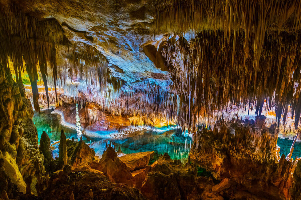Потолок пещеры украшен многочисленными сталактитами, которые свисают, словно ледяные сосульки. Освещение подчёркивает текстуру и формы каменных образований, создавая игру света и тени. Это место выглядит как настоящий природный шедевр, созданный временем и водой.