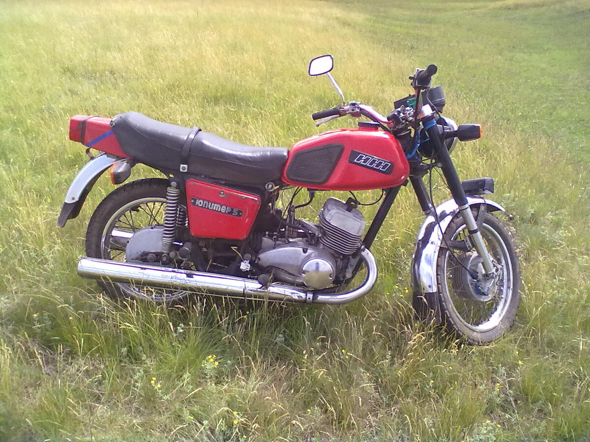 «Иж Юпитер 5» — Семейный работяга

Юпитер 5 – это более живой мотоцикл с неплохой приемистостью. По комфортабельности на голову выше своего оппонента. Двухцилиндровый двигатель мощнее и по скорости он быстрее. По паспорту скорость 125 км/час, но реально выжать и 150 только ремонт двигателя, потом обеспечен. Зажигание работает не стабильно и приносит много проблем. Расход топлива считается большим. Отличить легко по широкому двухместному седлу и двум амортизаторам сзади.

Концепция: Универсальный мотоцикл для двоих. «Юпитер» позиционировался как более практичный вариант, готовый перевозить водителя, пассажира и груз.

Главное визуальное отличие: седло и задняя часть

· Седло: Широкое, двухуровневое, полноценное двухместное. Пассажиру ехать значительно комфортнее.
· Задняя подвеска: Две пружинные стойки (два амортизатора) по бокам. Это классическая схема для большинства мотоциклов того времени.
· Задний щиток: Широкий, часто с интегрированными фарами-габаритами.

Плюсы «Юпитера 5»:

· Комфорт для двоих: Идеален для поездок с пассажиром.
· Более мягкая подвеска: Два амортизатора лучше справляются с неровностями, особенно под нагрузкой.
· Универсальность: На него проще установить грузовые кофры или прицеп.

Минусы:

· Больший вес: Чуть хуже разгонная динамика.
· «Раскачивание» на скорости: Из-за конструкции подвески может быть менее стабильным на высоких скоростях compared to «Планете».