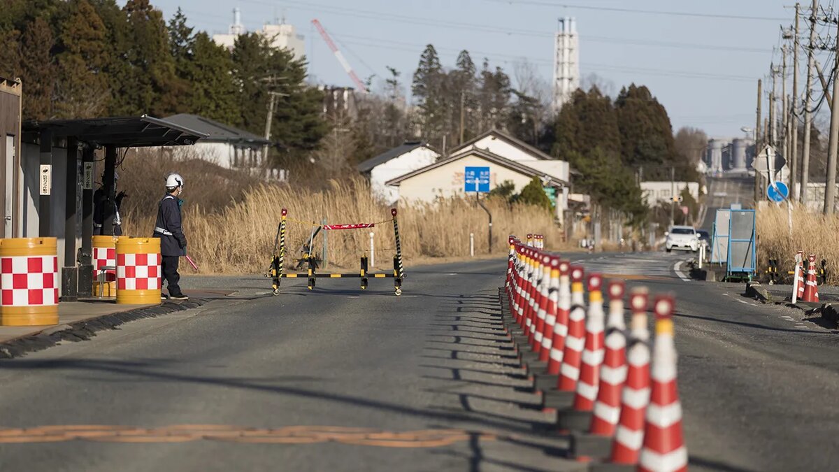   Hiro Komae / AP- Архивное фото. Сотрудники службы безопасности, на контрольно-пропускном пункте, на заднем плане которого видна часть атомной электростанции «Фукусима-1», в городе Окума