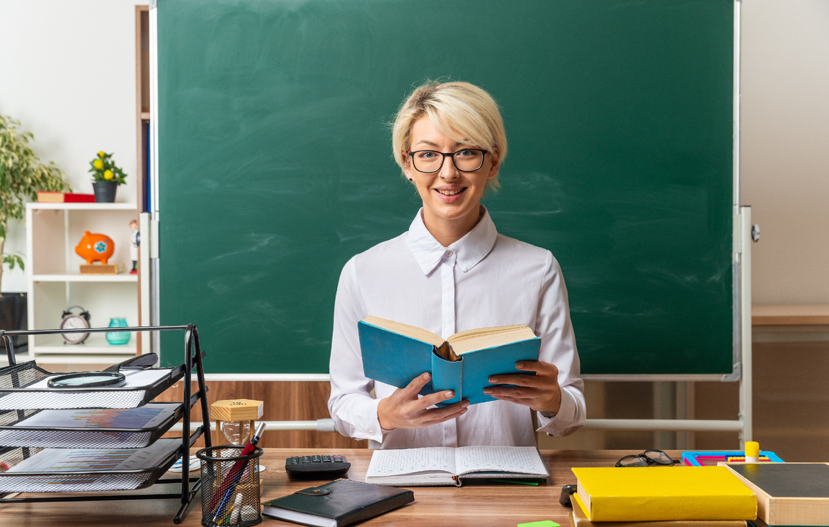 <a href="https://ru.freepik.com/free-photo/smiling-young-blonde-female-teacher-wearing-glasses-sitting-desk-with-school-supplies-classroom-holding-open-book-looking-front_17188348.htm">Изображение от stockking на Freepik</a>