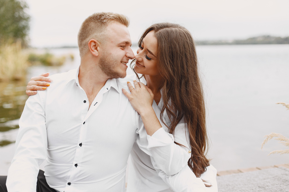 Влюблённая пара сидит у воды, смотрит друг на друга с улыбкой и выражает нежность, заботу и настоящую любовь.