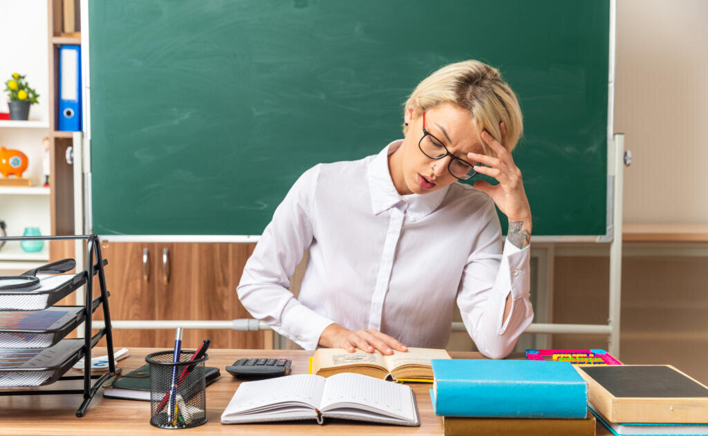    concentrated young blonde female teacher wearing glasses sitting at desk with school tools in classroom keeping hand on head and on open book reading book Журналист