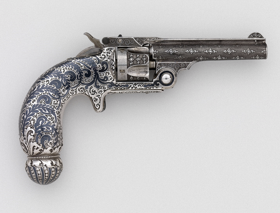 Револьвер Smith & Wesson калибра .32, одинарного действия
Производитель — Smith & Wesson (США, основана в 1852 году)
Украшение и отделка — Tiffany & Co. (основана в 1837 году, действует по настоящее время)
Дата: 1892–1893 годы
Происхождение: Спрингфилд (штат Массачусетс) и Нью-Йорк (штат Нью-Йорк)
Материалы: сталь, серебро, ниэль, никель.
Рукоять этого револьвера объединяет сразу три декоративные техники — рельефную чеканку (repoussé), травление и инкрустацию ниэлью.
Такое сочетание создало поразительно изысканный образ оружия, превращённого из утилитарного предмета в произведение ювелирного искусства.
Револьвер был представлен фирмой Smith & Wesson на Всемирной Колумбовой выставке в Чикаго в 1893 году, где служил примером того, как американское оружейное мастерство может сочетаться с роскошным художественным декором Tiffany & Co., став символом престижа, точности и ремесленного совершенства конца XIX века.
