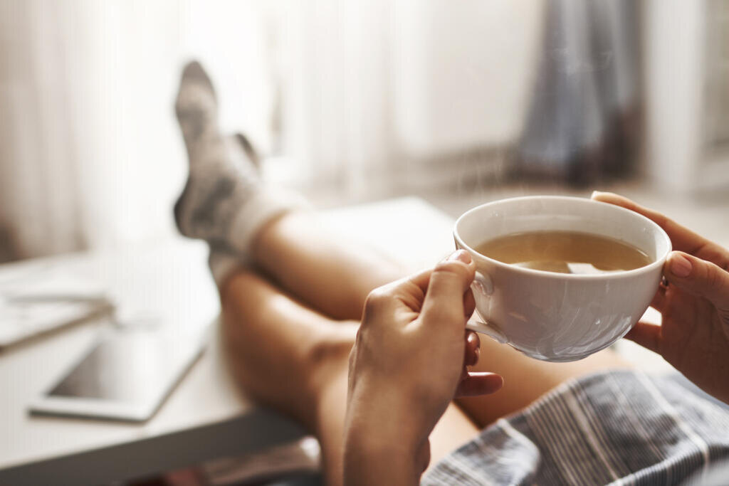    Cup of tea and chill. Woman lying on couch, holding legs on coffee table, drinking hot coffee and enjoying morning, being in dreamy and relaxed mood. Girl in oversized shirt takes break at home. Журналист