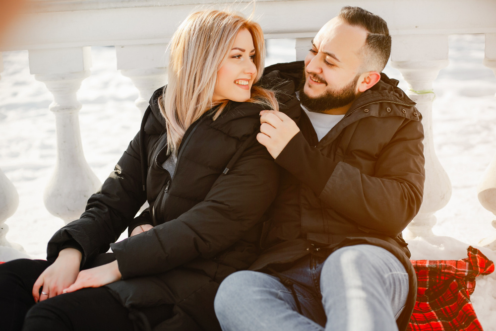Пара сидит вместе на улице зимой, улыбается и обнимается — момент тепла и любви в повседневной жизни.