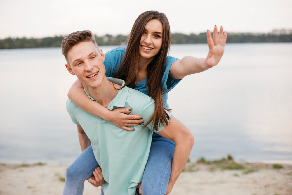 Девушка с улыбкой сидит на спине у парня на берегу озера, они счастливо смеются — символ лёгкости, близости и радости в отношениях.