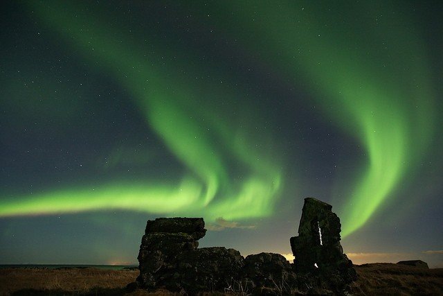 Над тёмным силуэтом старинных каменных руин танцуют изумрудные ленты северного сияния, разливая мягкое свечение по небу.