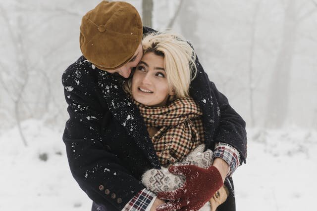 Мужчина обнимает женщину на снегу, и они улыбаются друг другу — символ тепла, доверия и нежной дружбы между мужчиной и женщиной.