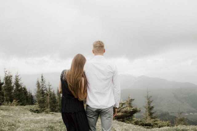 Пара стоит спиной на вершине горы, девушка обнимает мужчину за талию, перед ними туман и живописные горы