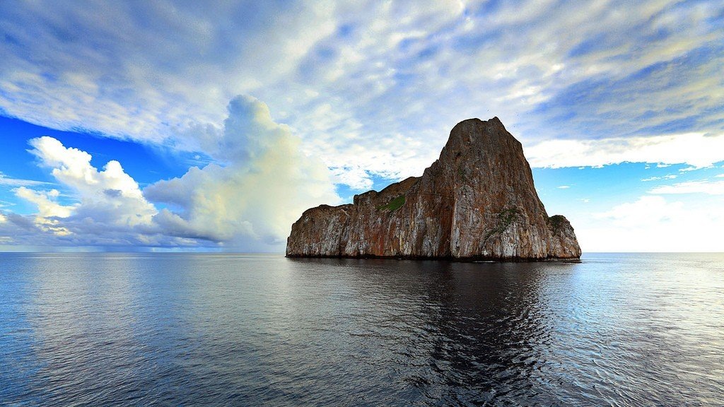 Одинокая скальная громада поднимается из спокойного Тихого океана, отражаясь в темной воде как природный монолит.