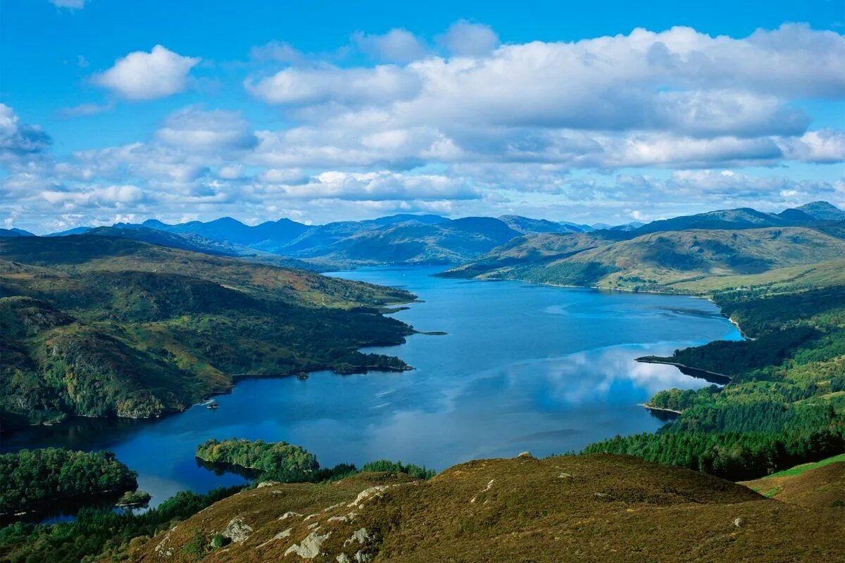 Шотландский пейзаж с изумрудными холмами и горами, глубоким синим озером, отражающим облака, и бескрайним горизонтом под ярким северным небом.