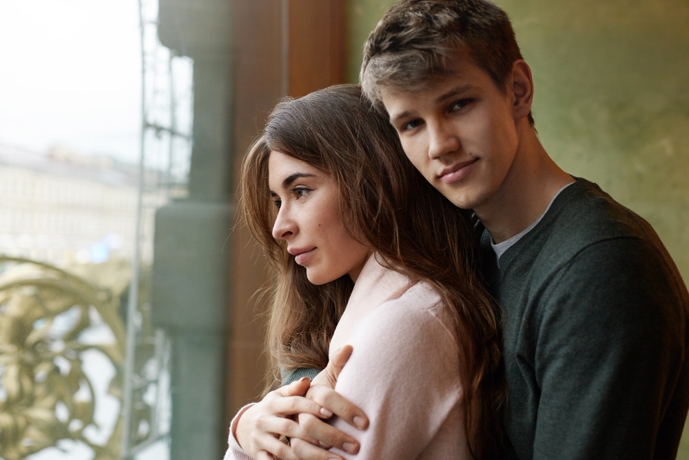 Мужчина обнимает женщину у окна — символ равновесия и поддержки в отношениях.