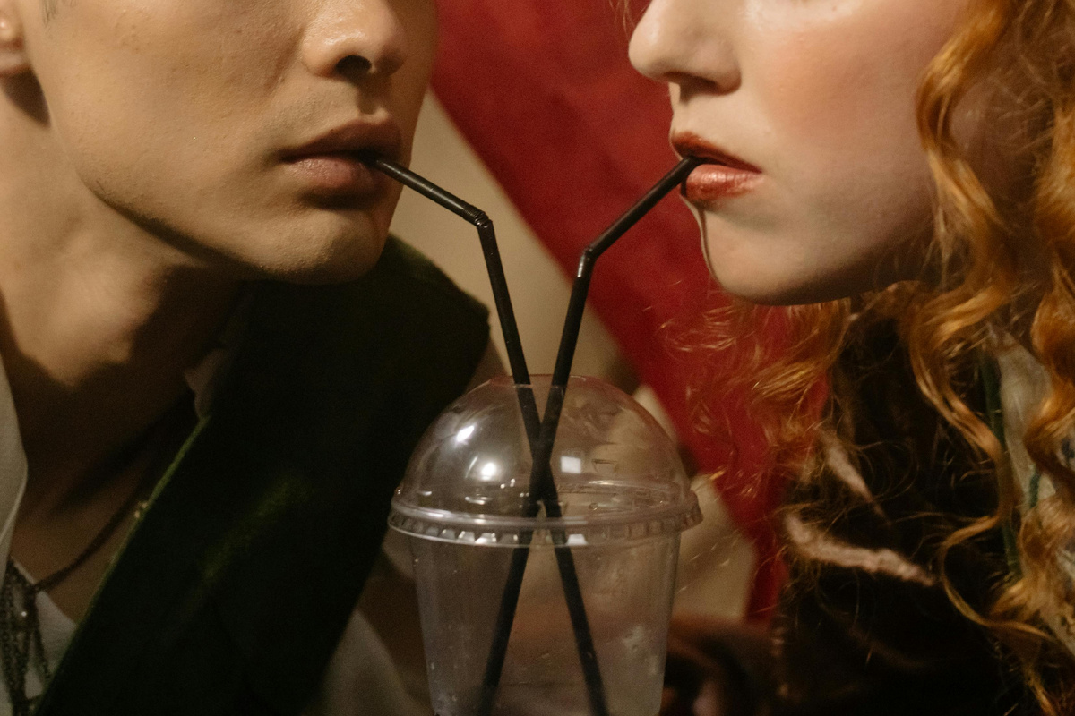 Молодая пара смотрит друг на друга, одновременно пьёт напиток через две трубочки — момент близости, страсти и соперничества, символизирующий тонкую грань между любовью и ревностью.