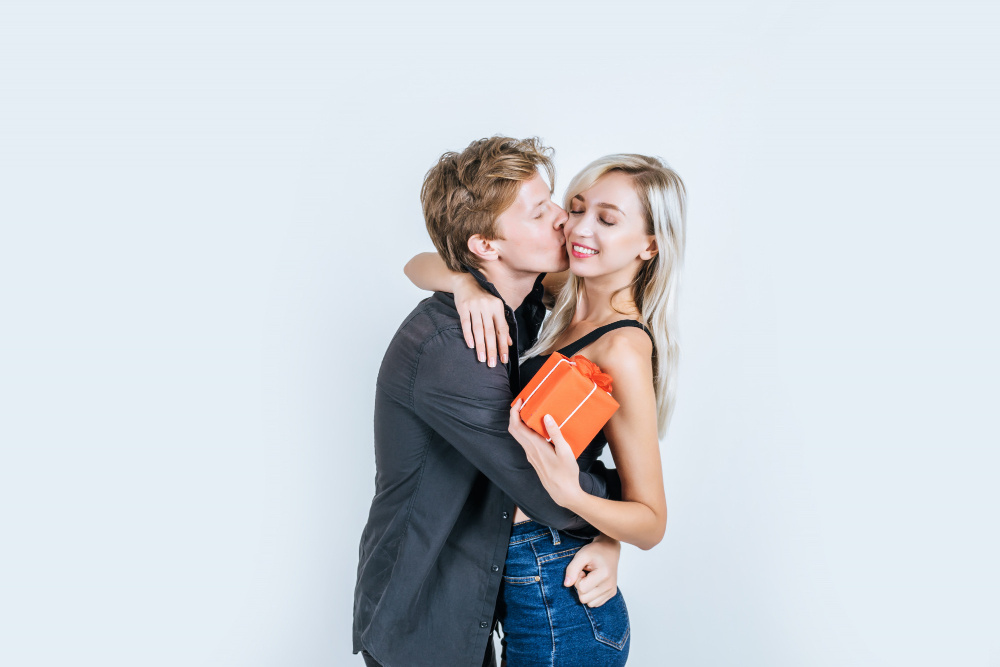 Молодой мужчина обнимает и целует девушку, а она улыбается, держа в руках книгу — символ доверия и гармонии в паре.