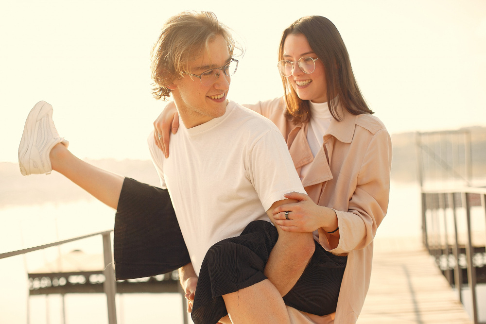 Молодая пара смеётся и играет на набережной, мужчина поднимает девушку на руках — момент радости и лёгкости в отношениях.