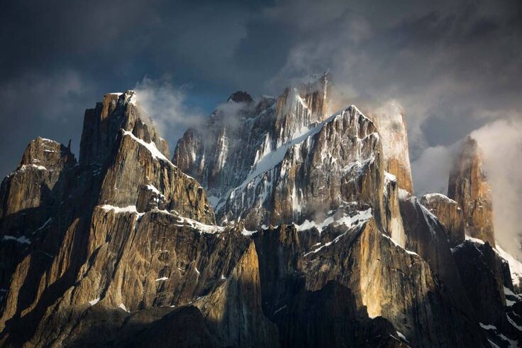 Зубчатые гранитные башни Каракорума поднимаются из тьмы, прорезая грозовые облака; резкий свет подчеркивает рельеф скал и тонкие полосы снега на карнизах.