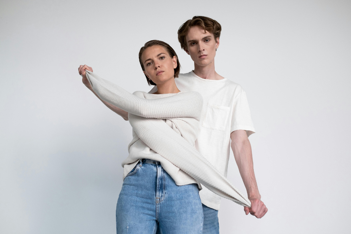 Пара стоит на светлом фоне, девушка в тёплом свитере, парень обнимает её сзади за рукав, спокойный момент близости