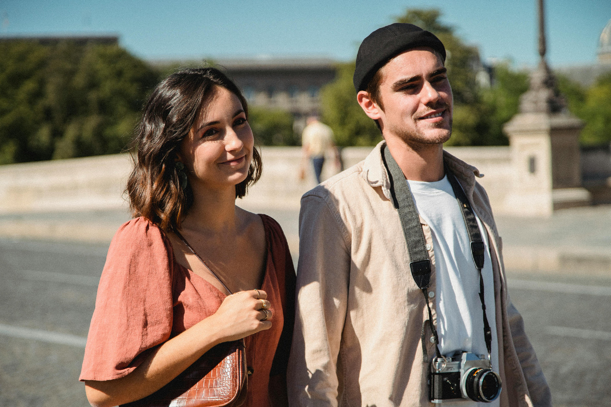 Молодая пара идёт по городу в солнечный день, мужчина с фотоаппаратом на шее, женщина улыбается — лёгкая атмосфера доверия и спокойствия.