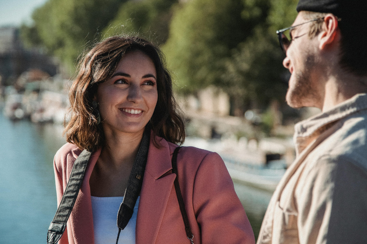 Молодая женщина с фотоаппаратом на шее улыбается мужчине во время прогулки у набережной, солнечный день и атмосфера лёгкости и доверия.
