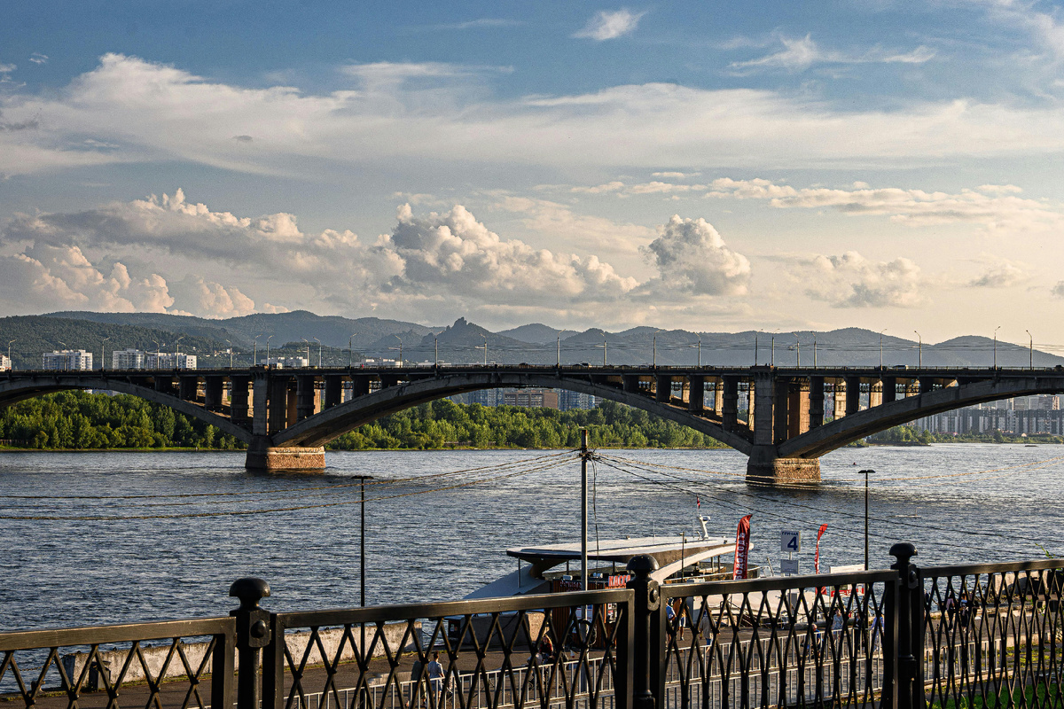 Вид на мост через реку Енисей в Красноярске в солнечный день, на заднем плане — горы и облака