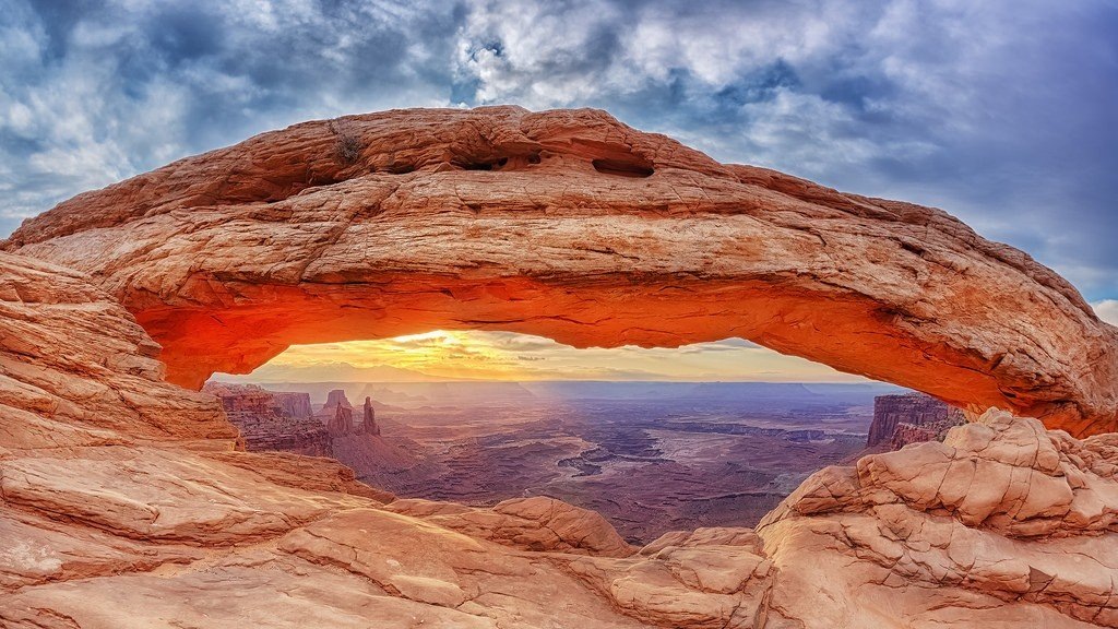 Закатное солнце, пробивающееся сквозь арку, заливает тёплым светом пустынный пейзаж, а на фоне видны далёкие каньоны и скальные образования. 