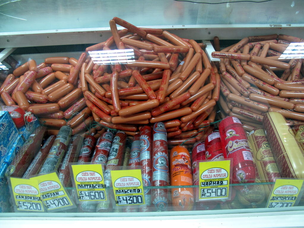 На заднем плане в холодильном прилавке бухты батончиков сосисок (по центру и справа) и сарделек (слева). На переднем плане батоны колбас (слева — полукопчёные, справа — варёные)