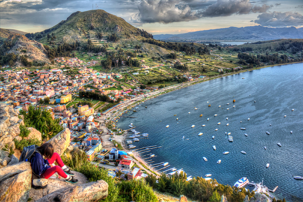 Путешественница сидит на скале и смотрит на бухту, город и горы под облачным небом.