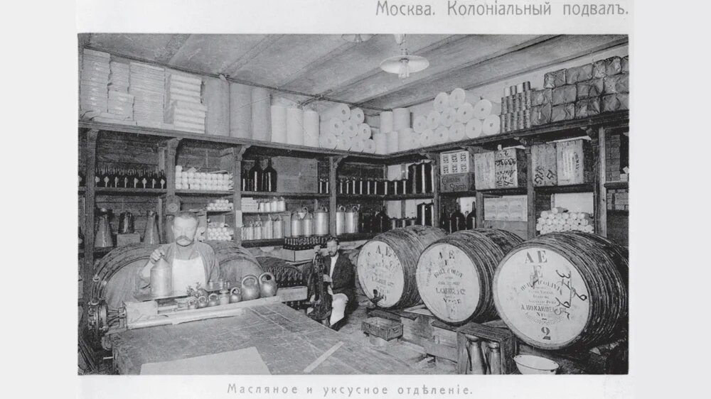 Историческая фотография московского «колониального подвала» начала XX века: масляное и уксусное отделение с бочками, стеклянными бутылями, мерной утварью и продавцами за рабочим столом. Атмосфера ранней торговли импортными товарами в России.