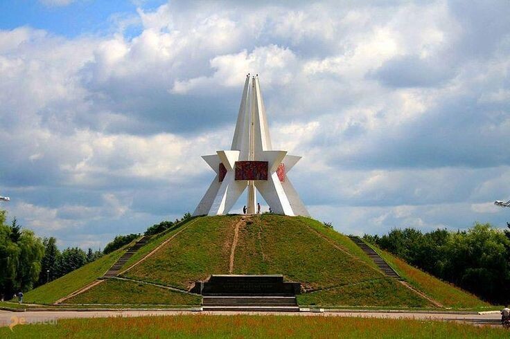 Монумент на Покровской горе в Брянске — белая звездообразная конструкция на вершине зелёного холма под облачным небом