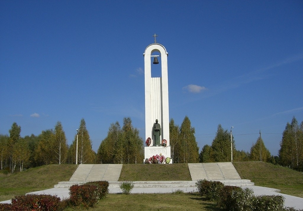 «Мемориальный комплекс „Хацунь“ — высокая белая арка с колоколом и памятником у подножия на фоне голубого неба и берёзовой рощи