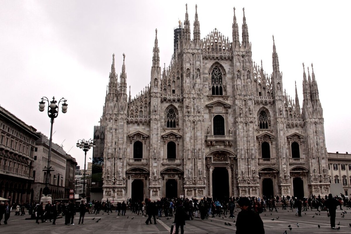 Неоготический Миланский собор на площади Дуомо в пасмурный день, люди гуляют по широкой площади перед фасадом.