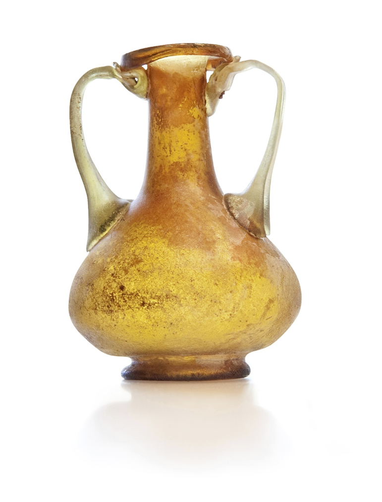 Римская стеклянная амфористическая фляга с двумя высокими прикладными ручками (I–III вв. н. э.). Такой тип часто называют двуручной флягой или мини-амфорой из стекла. Насыщенный янтарно-золотой цвет – очень ценился, потому что требовал контролируемого окисления железа при плавке.
