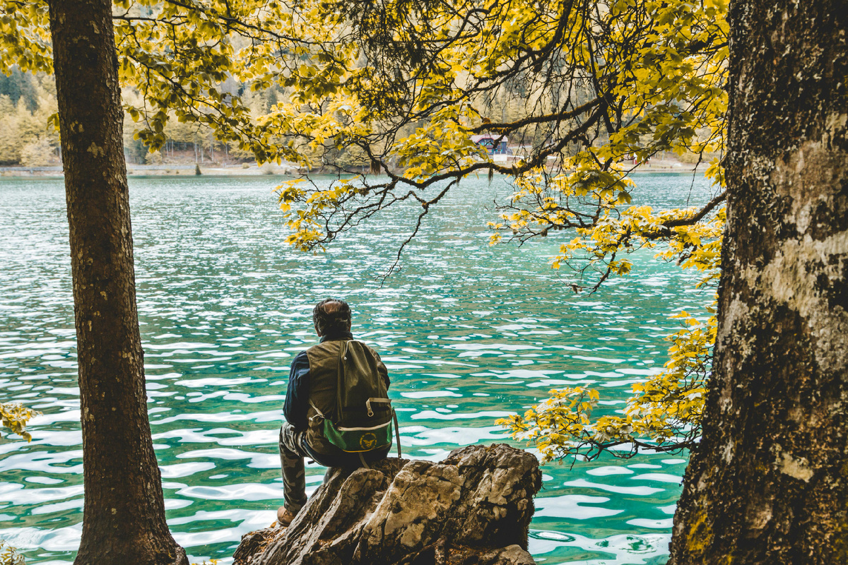 Путешественник с рюкзаком сидит на камне у бирюзового лесного озера среди деревьев