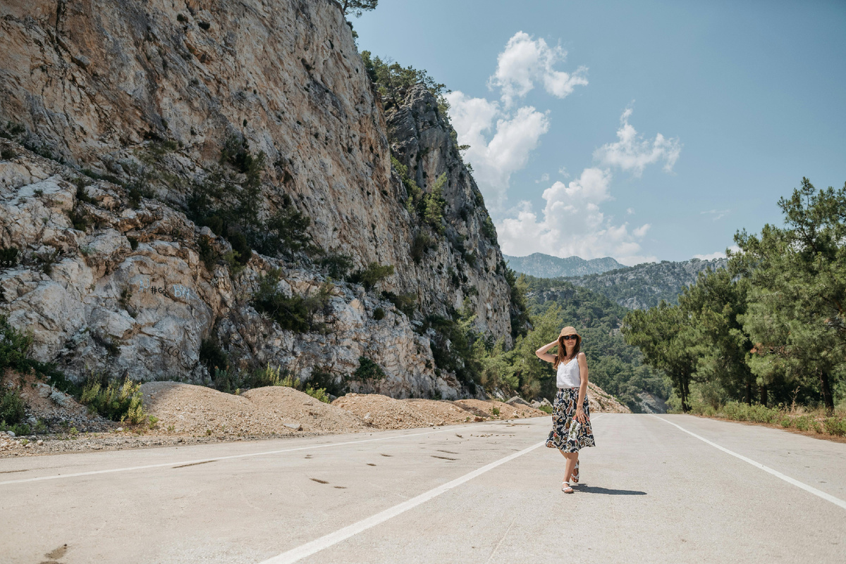 Женщина идёт по пустынной горной дороге на фоне скал и хвойных деревьев в солнечный день