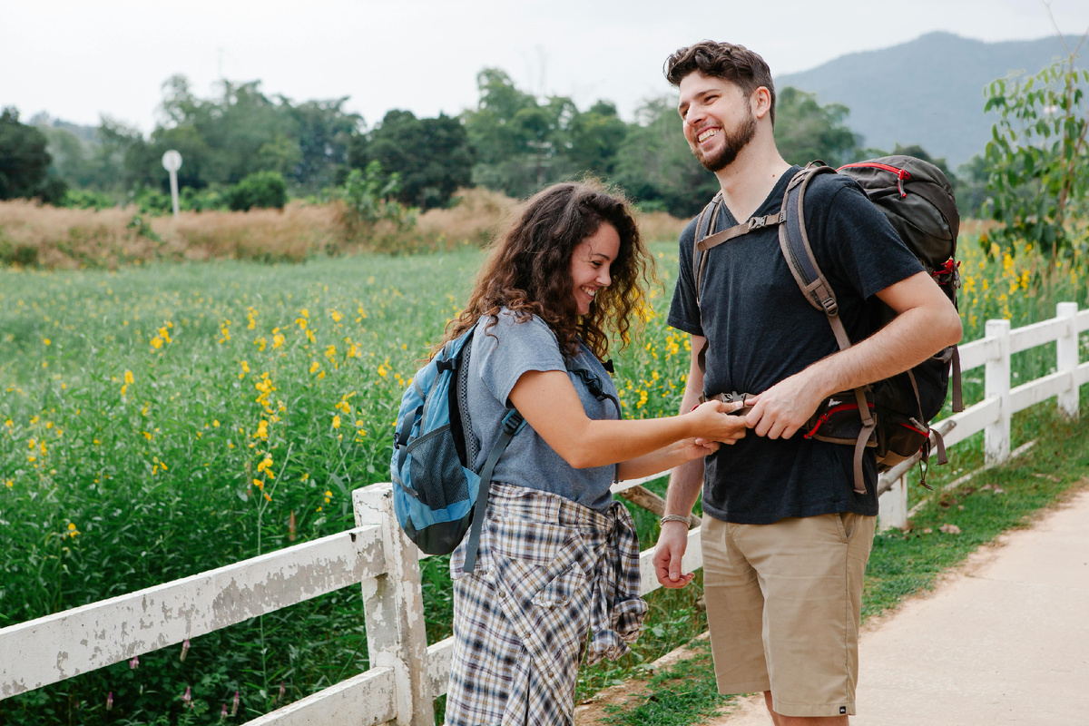Пара путешественников улыбается на тропе среди зелёных полей — лёгкий момент радости в походе.