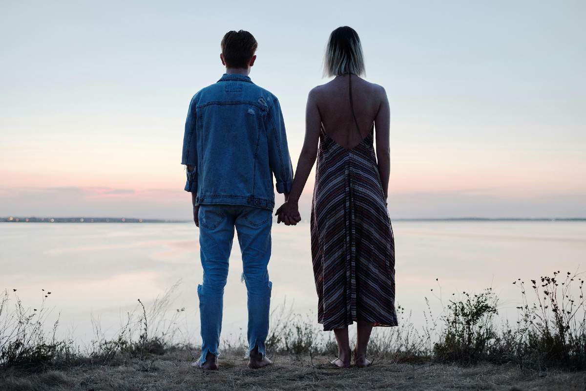 Пара стоит на берегу и держится за руки, смотря на спокойный закат над водой.
