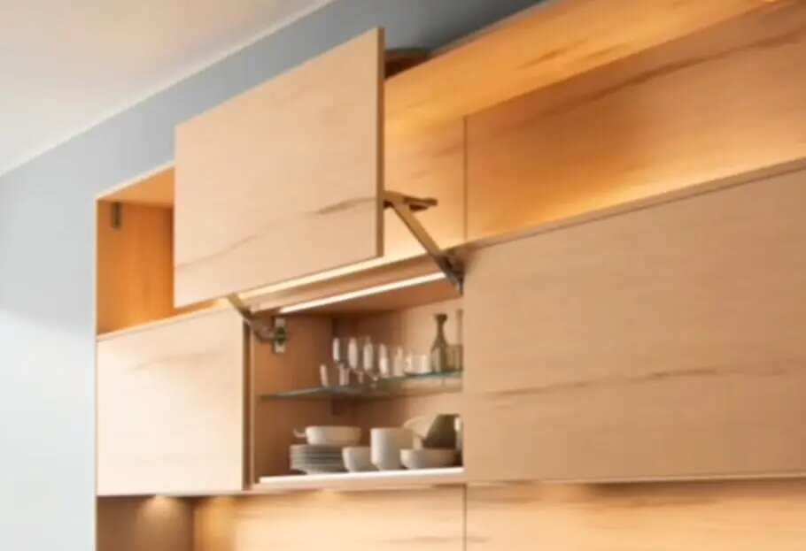 Верхний кухонный шкаф с современным подъемным механизмом. Фасады выполнены из светлого дерева, дверца открывается вверх, обеспечивая удобный доступ к посуде, расположенной на стеклянных полках внутри. Интегрированная подсветка подчеркивает аккуратный и функциональный дизайн кухни.