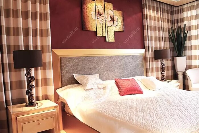 Спальня с большой кроватью, выполненной из массива с мягким изголовьем. Интерьер оформлен в тёплых тонах: бордовая акцентная стена, бежево-коричневые шторы и светлая деревянная мебель по бокам. Обстановка подчёркивает уют, комфорт и гармоничное сочетание натуральных материалов - идеальный фон для статьи о выборе кровати из массива.