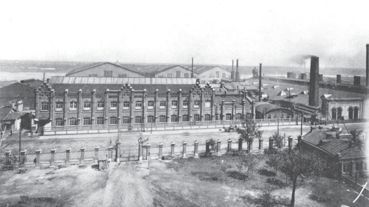 Вид части Невского судостроительного и механического завода от Шлиссельбургского проспекта в сторону Невы. На первом плане — здание кораблестроительной мастерской, за ним видны крыши и торцевые части большого и малых эллингов (слева направо). Начало XX века