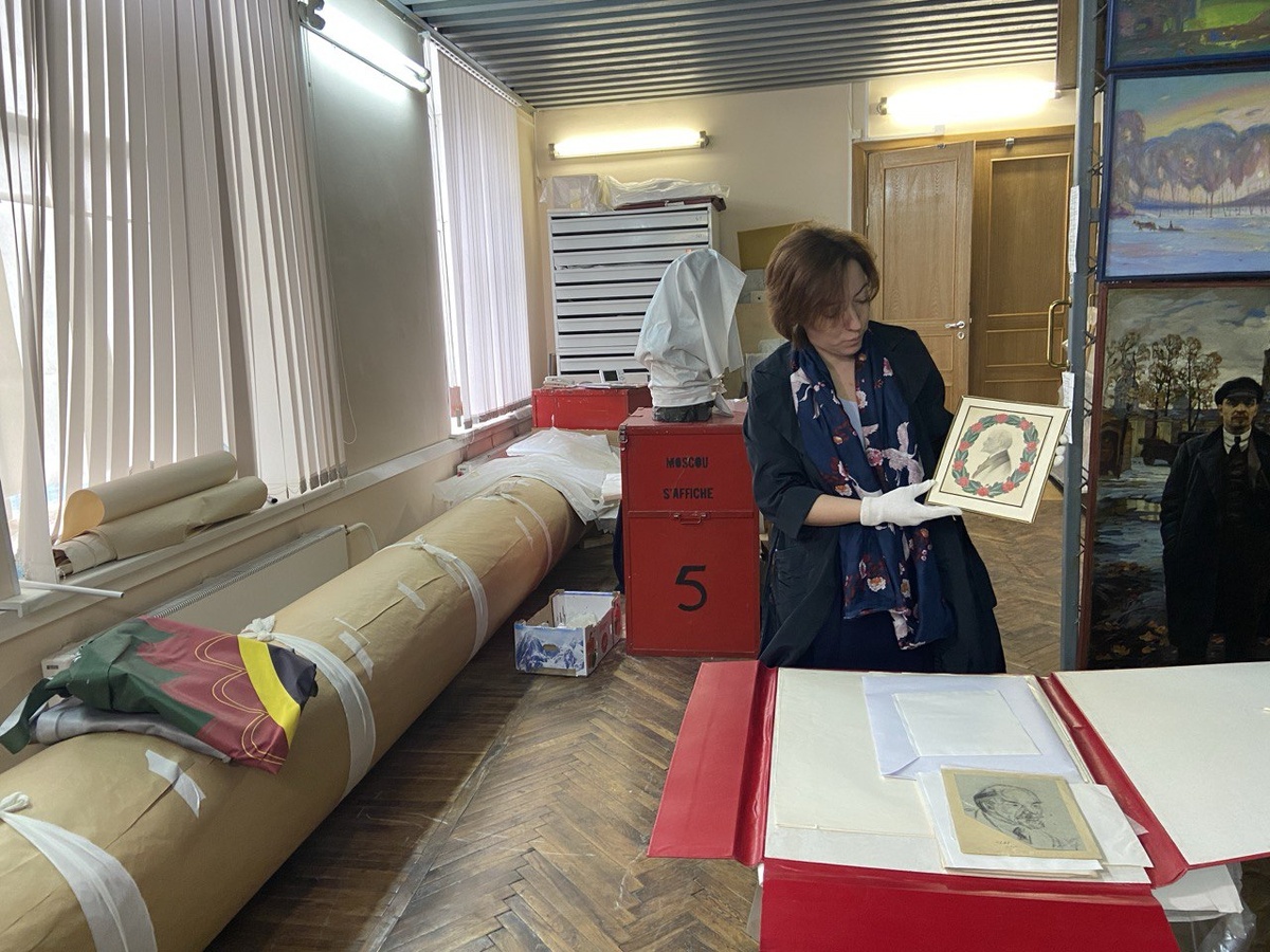 Хранитель фонда ИЗО и авторская графика ГМПИР Екатерина Костина показывает журналистам экспонат из коллекции графических изображений В. И. Ленина. В руках – работа художника по фарфору Сергея Чехонина, в папке на столе – рисунок известного авангардиста Натана Альтмана. Фото: пресс-служба ГМПИР, апрель 2025 г. 