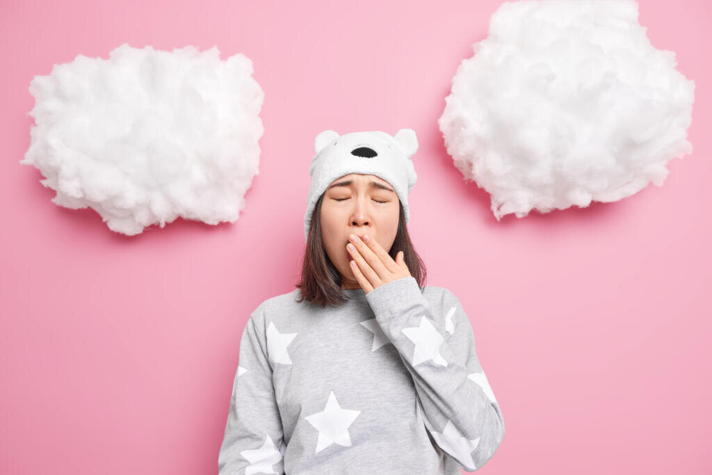    Sleepy teenage Asian girl covers mouth yawns wants to sleep has problem of insomnia dressed in nightwear soft bear hat isolated over pink background. Bedtime energy charge concept. Lack of vivacity Журналист