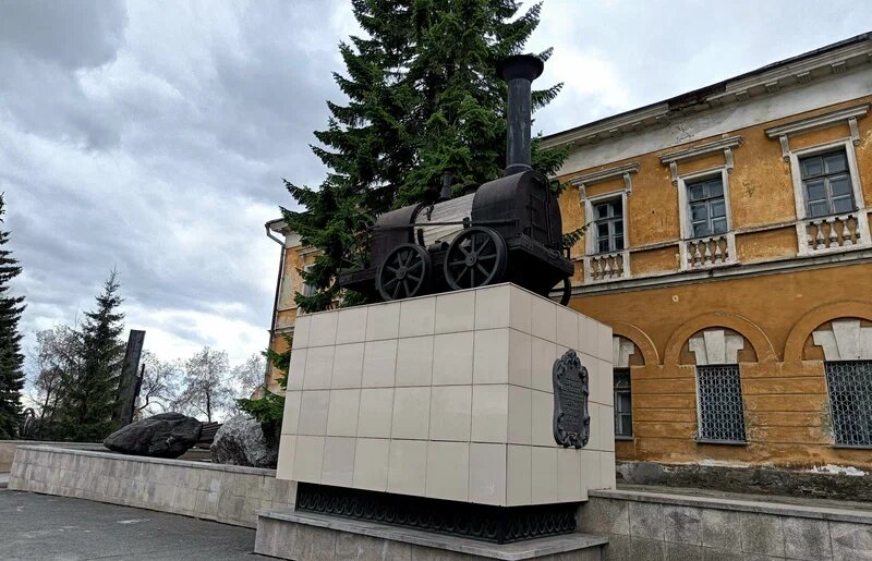Памятник паровозу Черепановых на фоне заводских цехов в Нижнем Тагиле, узкие рельсы уходят к старой шахте