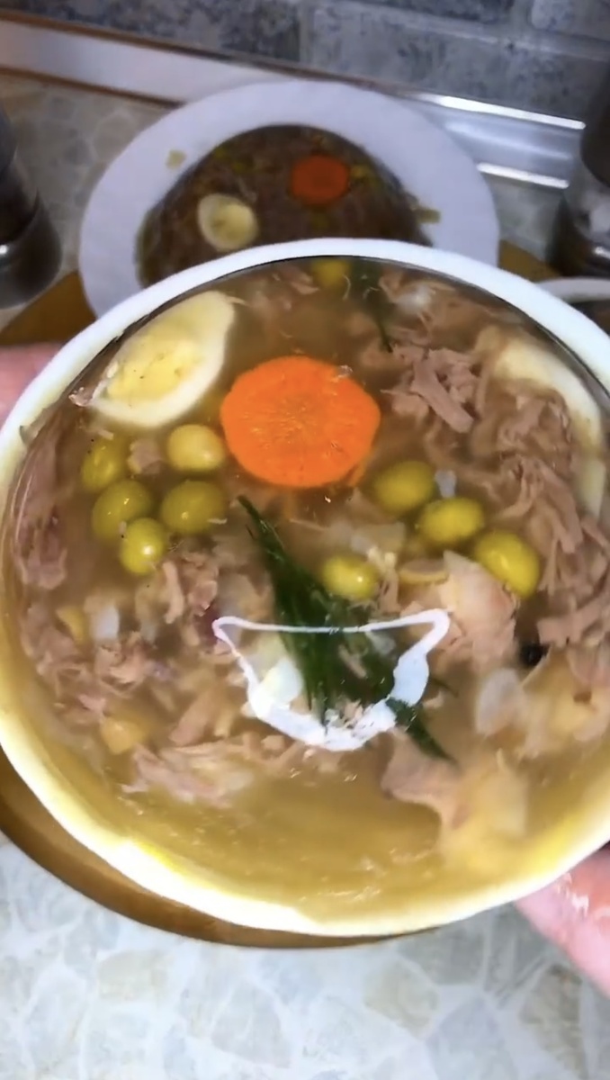 Ингредиенты:
Говяжья голяшка или рулька (на кости): 1,5–2 кг (для хорошего застывания)
Вода: примерно 2 л воды на 1 кг мяса
Морковь: 2-3 шт.
Лук репчатый: 1-2 шт.
Чеснок: 5-7 зубчиков
Лавровый лист: 3-4 шт.
Черный перец горошком: 10-15 шт.
Соль: по вкусу
Зелень (петрушка, укроп): для украшения 
Пошаговая инструкция:
Подготовка мяса: Мясо и кости тщательно промойте. Желательно вымочить их в холодной воде в течение нескольких часов (можно на ночь), чтобы удалить лишнюю кровь и сделать бульон прозрачнее.
Первичная варка: Залейте мясо холодной водой и доведите до кипения. Как только вода закипит, слейте ее, а мясо промойте. Это поможет избавиться от пены и получить чистый бульон.
Основная варка бульона: Залейте мясо свежей холодной водой (в соотношении примерно 1 кг мяса к 2 л воды). Доведите до кипения, затем максимально уменьшите огонь. Бульон должен не кипеть, а медленно томиться под приоткрытой крышкой в течение 6-8 часов. В процессе снимайте образующуюся пену и жир.
Добавление овощей и специй: Примерно за 2-3 часа до окончания варки добавьте целую очищенную морковь, неочищенную луковицу (для цвета), лавровый лист и перец горошком. Посолите по вкусу.
Извлечение и разделка: Аккуратно достаньте из бульона мясо, овощи и специи. Морковь можно нарезать фигурными кусочками для украшения. Мясо отделите от костей и разберите на мелкие волокна или нарежьте кубиками. Чеснок пропустите через пресс и добавьте к мясу.
Подготовка форм: Разложите мясо с чесноком по формочкам или глубоким тарелкам. Сверху красиво уложите кусочки моркови и веточки свежей зелени.
Заливка и застывание: Процедите бульон через мелкое сито или марлю, чтобы он был идеально прозрачным. Залейте бульоном содержимое формочек.
Охлаждение: Дайте холодцу остыть до комнатной температуры, затем уберите в холодильник до полного застывания (обычно требуется несколько часов или ночь). 