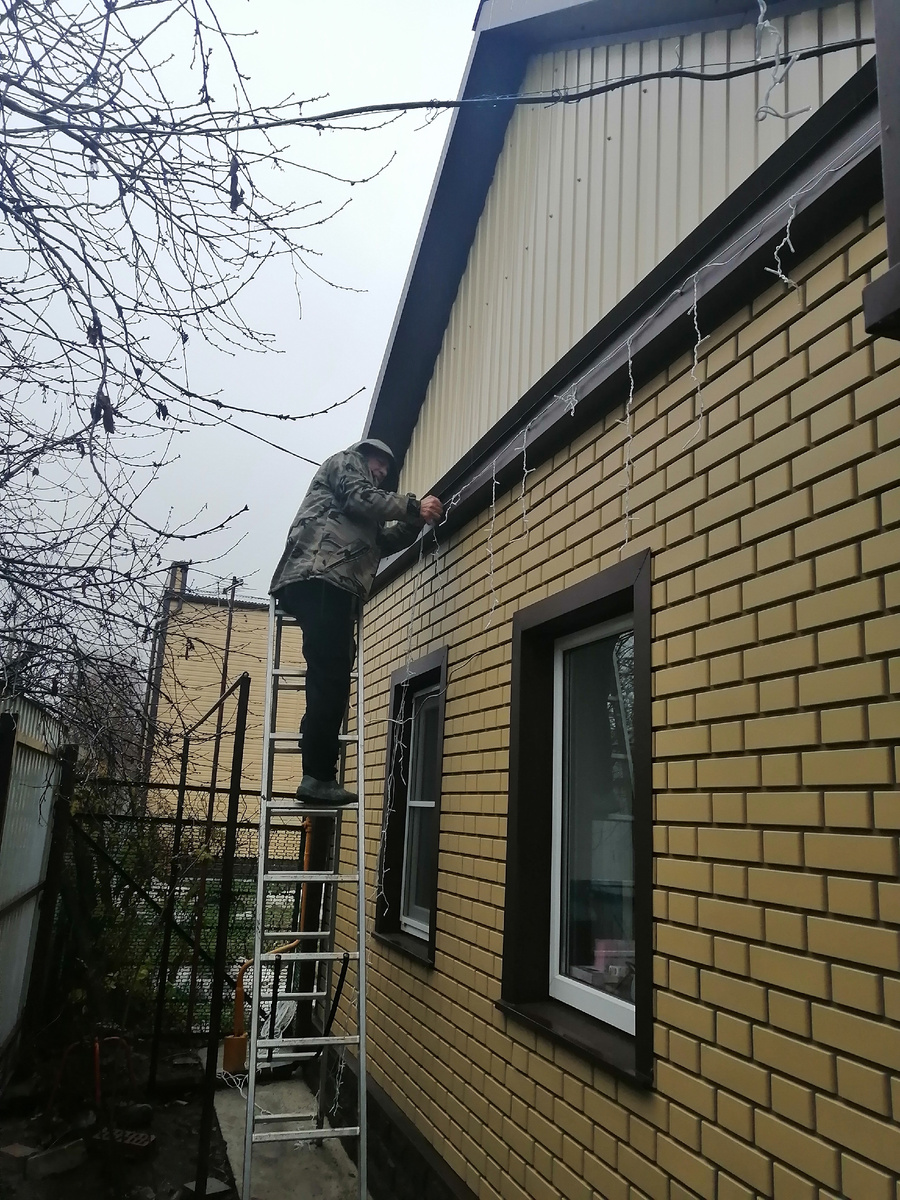 Льет дождь ледяной , но муж стойко вешает гирлянду.