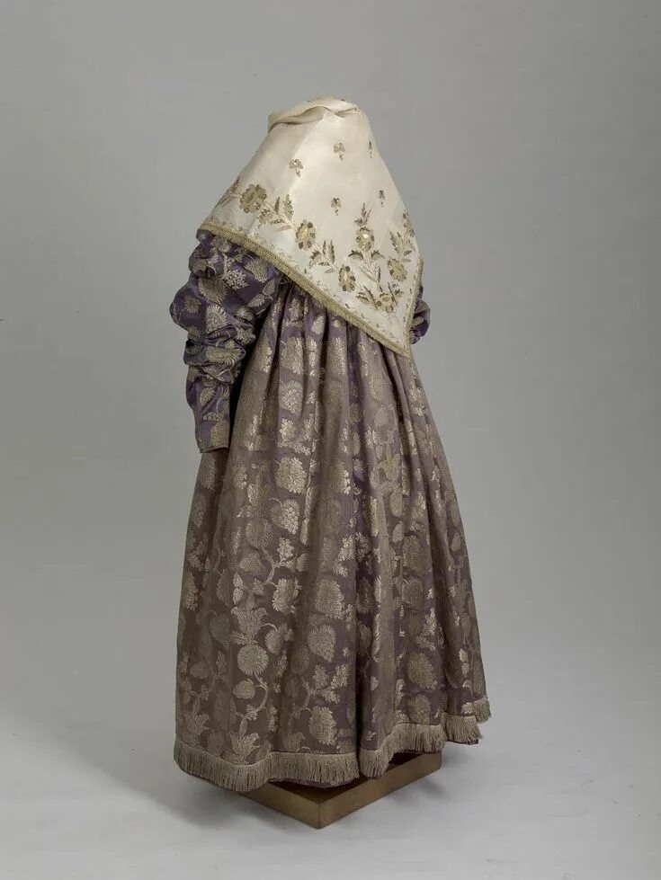 На изображении показан сарафан — ключевой элемент сарафанного комплекса. В Костромской губернии преобладали туникообразные, прямые и косоклинные модели. Сарафаны украшали вышивкой, лентами, тесьмой и бахромой. Праздничные варианты шили из шёлка, парчи или полупарчи. 