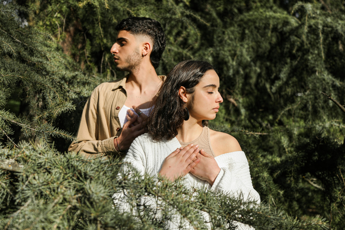 Парень и девушка стоят среди хвойных веток, смотрят в разные стороны; у девушки руки на груди, спокойная задумчивая атмосфера.