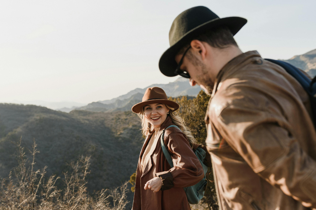 Пара туристов в шляпах идёт по горной тропе на фоне холмов и закатного света